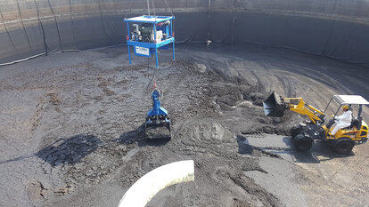 Ansicht von oben, Vogelperspektive, eine Tanks mit schlammartigem Inhalt, welcher durch einen kleinen gelben Bagger, rechts im Bild, zusammen geschoben wird. Ein königsblauer Greifarm, in der Mitte des Bildes, heb beziehungsweise lädt die schlammartige Masse mit seinem Greifer auf..