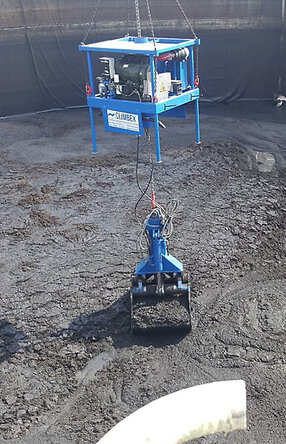 Ansicht von oben, Vogelperspektive, eine Tanks mit schlammartigem Inhalt, welcher durch einen kleinen gelben Bagger, rechts im Bild, zusammen geschoben wird. Ein königsblauer Greifarm, in der Mitte des Bildes, heb beziehungsweise lädt die schlammartige Masse mit seinem Greifer auf..