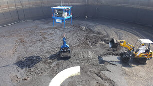 Ansicht von oben, Vogelperspektive, eine Tanks mit schlammartigem Inhalt, welcher durch einen kleinen gelben Bagger, rechts im Bild, zusammen geschoben wird. Ein königsblauer Greifarm, in der Mitte des Bildes, heb beziehungsweise lädt die schlammartige Masse mit seinem Greifer auf..