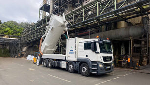 Weißer LKW mit Climbex Logos und einem gekippten Tank hinten drauf steht vor einem Fabrikgebäude, außen verlaufen Metallstützen. Hinten links neben dem LKW steht ein Mitarbeiter in weißem Anzug und mit orangefarbenen Arbeitshelm, er steuert den Tank. Hinter dem LKW und Mitarbeiter ist ein Gabelstapler zu sehen.