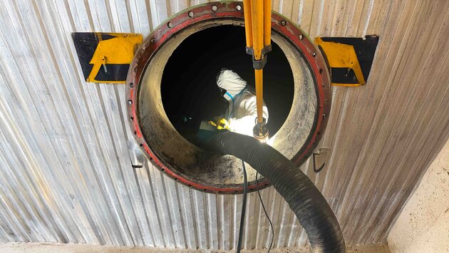 Das Foto zeigt einen von außen Fotografierten Tank mit einer runden Öffnung, im inneren des Tanks steht ein Arbeiter mit Schutzanzug und Maske während er ein Rohr, das von außen kommt, festhält.