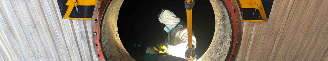 Das Foto zeigt einen von außen Fotografierten Tank mit einer runden Öffnung, im inneren des Tanks steht ein Arbeiter mit Schutzanzug und Maske während er ein Rohr, das von außen kommt, festhält.