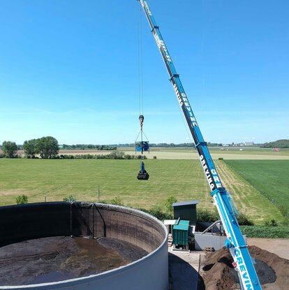Ein Mitarbeiter sitzt mit Schutzanzug, Gasmaske & Handschuhen auf einem Mini-Bagger der vorne einen Aufsatz zum zusammenschieben von Materalien hat. Er arbeitet im inneren eines Tanks und schieb eine Schlamm ähnliche Substanz rum.