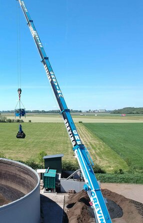 Ein Mitarbeiter sitzt mit Schutzanzug, Gasmaske & Handschuhen auf einem Mini-Bagger der vorne einen Aufsatz zum zusammenschieben von Materalien hat. Er arbeitet im inneren eines Tanks und schieb eine Schlamm ähnliche Substanz rum.