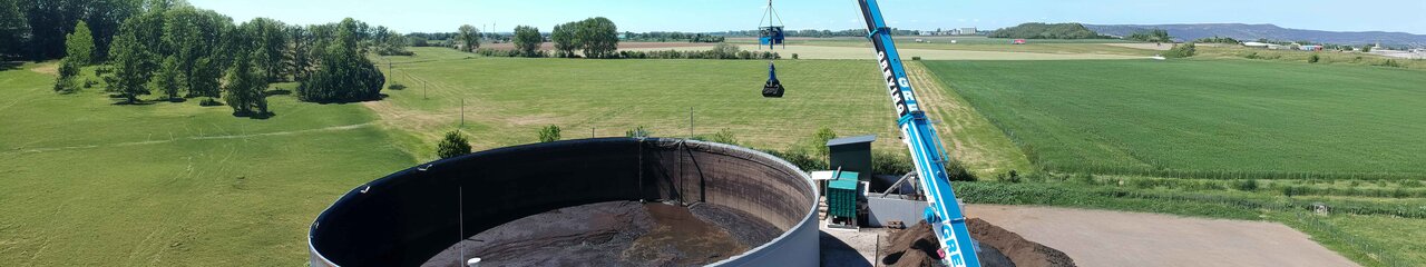 Ein Mitarbeiter sitzt mit Schutzanzug, Gasmaske & Handschuhen auf einem Mini-Bagger der vorne einen Aufsatz zum zusammenschieben von Materalien hat. Er arbeitet im inneren eines Tanks und schieb eine Schlamm ähnliche Substanz rum.