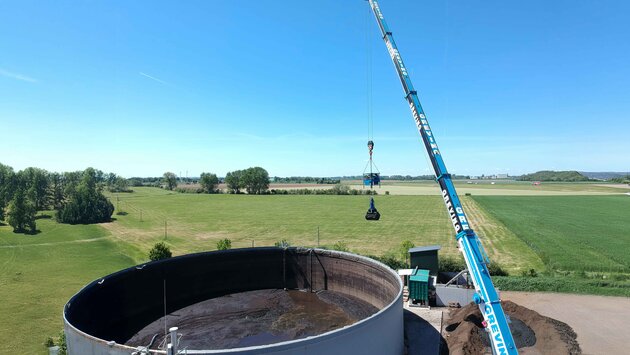 Ein Mitarbeiter sitzt mit Schutzanzug, Gasmaske & Handschuhen auf einem Mini-Bagger der vorne einen Aufsatz zum zusammenschieben von Materalien hat. Er arbeitet im inneren eines Tanks und schieb eine Schlamm ähnliche Substanz rum.