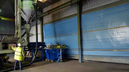 Zu sehen ist eine Halle mit einer relativ großen Siebanlage die von einem Mitarbeiter in neongelber Arbeitskleidung und Schutzhelm bedient wird.