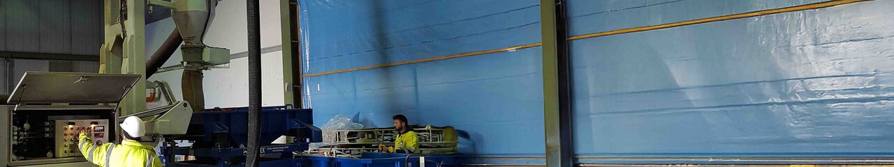 Zu sehen ist eine Halle mit einer relativ großen Siebanlage die von einem Mitarbeiter in neongelber Arbeitskleidung und Schutzhelm bedient wird.