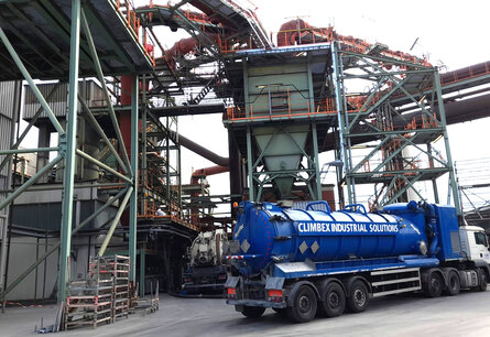 Zu sehen ist das Äußere eines Fabrikgeländes mit diversen Rohren und Metallgeländern, außerdem steht darin eine trichterförmige Anlage. Vor der Anlage steht ein LKW mit einem großen Tank.