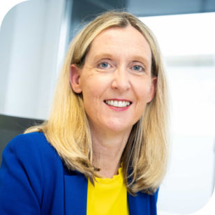 Eine Mitarbeiterin mittleren alters mit blonden Haaren lächelt, leicht zur Kamera gedreht, in die Kamera. Sie trägt einen königsblauen Blazer mit einem gelbem T-Shirt drunter und am Schreibtisch sitzend.