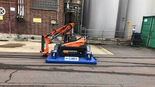 Roboter, orangefarben, mit gesenktem Greifarm stehend auf einer königsblauen Platte die über ein Schienensystem auf einem Betriebshof fährt. Im Hintergrund sind eine Backstein-Fassade und Silos oder Tanks zu sehen.