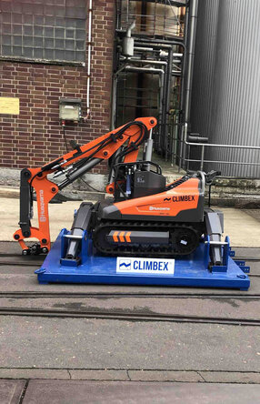 Roboter, orangefarben, mit gesenktem Greifarm stehend auf einer königsblauen Platte die über ein Schienensystem auf einem Betriebshof fährt. Im Hintergrund sind eine Backstein-Fassade und Silos oder Tanks zu sehen.