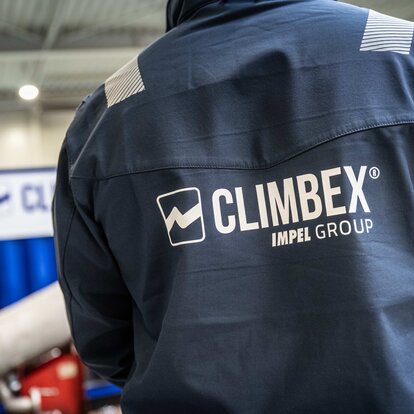 Aufnahme eines Rückens eines Climbex Mitarbeiter, welcher eine dunkelblaue Arbeiter-Jacke trägt auf der das Climbex Logos mit dem Zusatz 'Impel Group' Schriftzug zu sehen ist. Im Hintergrund sind unscharf ein Gerät und Climbex-Container zu sehen.