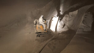 Zu sehen ist ein Arbeiter in Schutzanzug im inneren eines Silos auf einem Bagger ähnliche Fahrzeug an dem ein Schlauch befestigt ist, womit ein Pulver aufgesaugt wird.