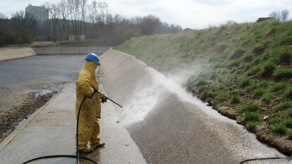 Es steh eine Person mit gelbem Ganzkörperanzug und königsblauen Arbeitshelm in einer offenen, Kanal ähnlichen, Grube und hält dabei einen Hochdruckstrahler auf die Innenwand.
