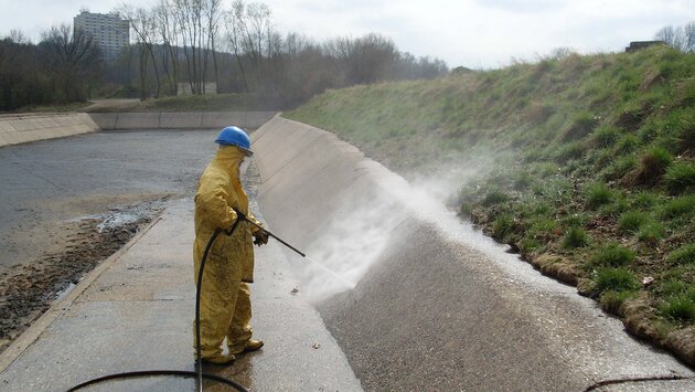 Es steh eine Person mit gelbem Ganzkörperanzug und königsblauen Arbeitshelm in einer offenen, Kanal ähnlichen, Grube und hält dabei einen Hochdruckstrahler auf die Innenwand.