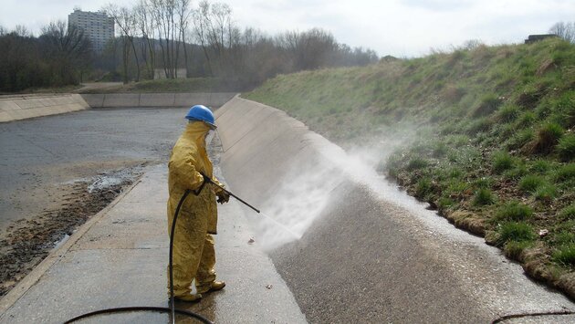 Es steh eine Person mit gelbem Ganzkörperanzug und königsblauen Arbeitshelm in einer offenen, Kanal ähnlichen, Grube und hält dabei einen Hochdruckstrahler auf die Innenwand.