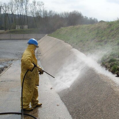 Es steh eine Person mit gelbem Ganzkörperanzug und königsblauen Arbeitshelm in einer offenen, Kanal ähnlichen, Grube und hält dabei einen Hochdruckstrahler auf die Innenwand.