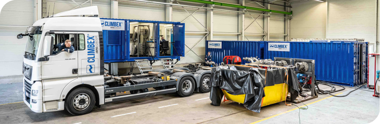 Halle von innen, darin steht einen Firmen-LKW mit Arbeiter im Fahrerhaus, dazu sind verschiedenen Firmen-Containern und andere Geräte zu sehen.