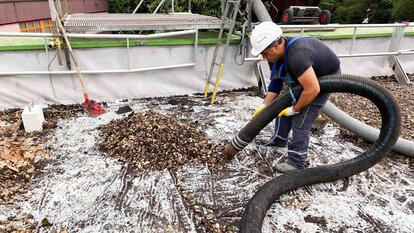 Zu sehen ist ein Arbeiter in Arbeitskleidung und mit Schutzhelm, der mit einem Rohr zusammengeschobenes Kies von einen Dach aufsaugt.