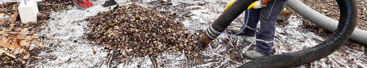 Zu sehen ist ein Arbeiter in Arbeitskleidung und mit Schutzhelm, der mit einem Rohr zusammengeschobenes Kies von einen Dach aufsaugt.