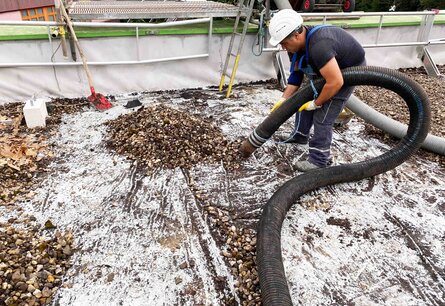 Zu sehen ist ein Arbeiter in Arbeitskleidung und mit Schutzhelm, der mit einem Rohr zusammengeschobenes Kies von einen Dach aufsaugt.