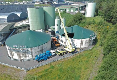 Aufnahme aus der Vogelperspektive, eines grünlichen Fabrikgebäudes, welches aus zwei großen flachen Silos im Vordergrund und zwei schmalen hohen Silos im Hintergrund besteht. Vor und zwischen den Silos stehen unterschiedliche LKWs, einer mit Tank, einer mit offener beladener Ladefläche und einer mit einem Kran der gerade einen Maschine in einen der Tanks hebt. Im Hintergrund sind weitere Fabriken sowieso Wiesen und Wälder zu sehen.