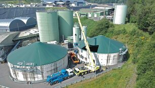 Aufnahme aus der Vogelperspektive, eines grünlichen Fabrikgebäudes, welches aus zwei großen flachen Silos im Vordergrund und zwei schmalen hohen Silos im Hintergrund besteht. Vor und zwischen den Silos stehen unterschiedliche LKWs, einer mit Tank, einer mit offener beladener Ladefläche und einer mit einem Kran der gerade einen Maschine in einen der Tanks hebt. Im Hintergrund sind weitere Fabriken sowieso Wiesen und Wälder zu sehen.