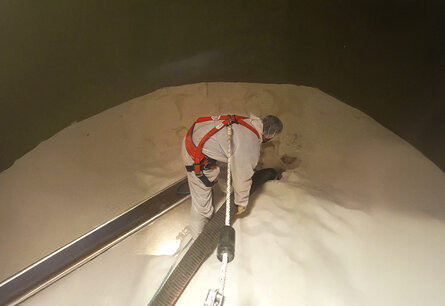 Ein Mitarbeiter in weißem Ganzkörperanzug, Haarnetz und einem roten Geschirr, das an dem ein Seil befestigt ist, steht mittig in einem Tank auf weißem Pulver. Der Mitarbeiter hält mit beiden Händen ein Rohr womit es das Pulver vermutlich aufsaugt, links neben ihm ist ein schwarze Brett um aus dem Tank ein- oder auszusteigen.