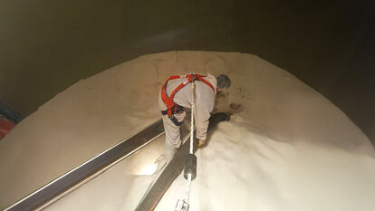 Ein Mitarbeiter in weißem Ganzkörperanzug, Haarnetz und einem roten Geschirr, das an dem ein Seil befestigt ist, steht mittig in einem Tank auf weißem Pulver. Der Mitarbeiter hält mit beiden Händen ein Rohr womit es das Pulver vermutlich aufsaugt, links neben ihm ist ein schwarze Brett um aus dem Tank ein- oder auszusteigen.