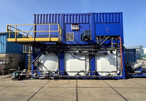 Zu sehen ist eine Konstruktion auf einem Gelände, bestehend aus drei großen weißen Tanks auf denen jeweils ein königsblauer Container steht in dem vermutlich die Motoren und Rohre sind.