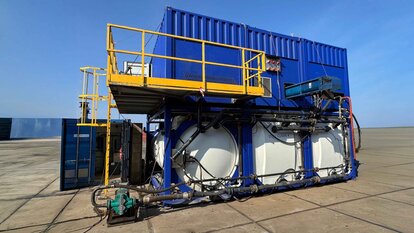 Zu sehen ist eine Konstruktion auf einem Gelände, bestehend aus drei großen weißen Tanks auf denen jeweils ein königsblauer Container steht in dem vermutlich die Motoren und Rohre sind.