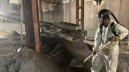 Halle mit einem Haufen an Schutt, darin stehend ein Mitarbeiter in Schutzanzug und Maske mit einer Schaufel, der Schutt auf einen Kasten auf einem Förderband auflädt. Das Förderband hängt zwischen Metallpfeilern und läuft nach hinten aus dem Bild raus.