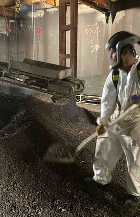 Halle mit einem Haufen an Schutt, darin stehend ein Mitarbeiter in Schutzanzug und Maske mit einer Schaufel, der Schutt auf einen Kasten auf einem Förderband auflädt. Das Förderband hängt zwischen Metallpfeilern und läuft nach hinten aus dem Bild raus.