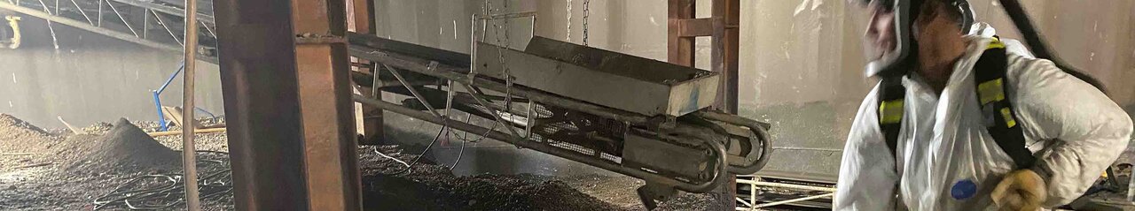 Halle mit einem Haufen an Schutt, darin stehend ein Mitarbeiter in Schutzanzug und Maske mit einer Schaufel, der Schutt auf einen Kasten auf einem Förderband auflädt. Das Förderband hängt zwischen Metallpfeilern und läuft nach hinten aus dem Bild raus.