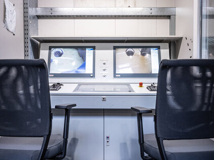 Frontalter Blick auf zwei Stühle, links und rechts an den Ecken des Bildes, vor einem Kontrollpult mit Monitoren.