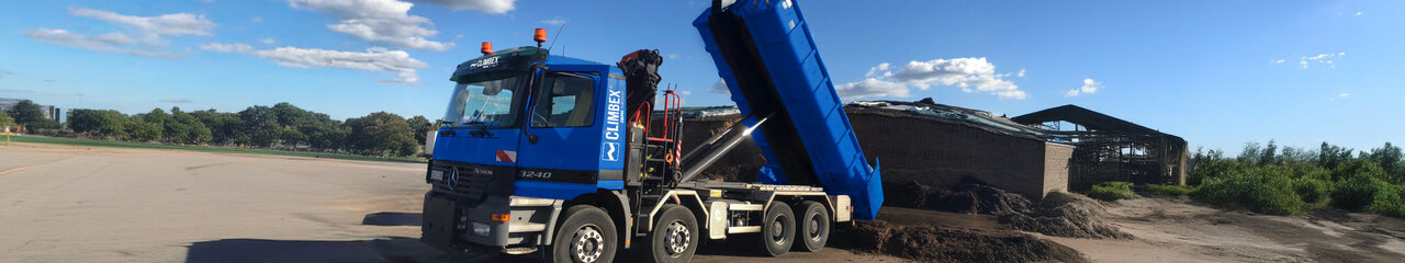 Zu sehen ist ein LKW, in der Mitte des Bildes, mit gekippter Ladefläche wie er Dreck ablädt. Hinter dem LKW steht eine Halle vor der weiterer Dreck liegt.