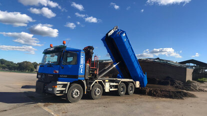 Zu sehen ist ein LKW, in der Mitte des Bildes, mit gekippter Ladefläche wie er Dreck ablädt. Hinter dem LKW steht eine Halle vor der weiterer Dreck liegt.