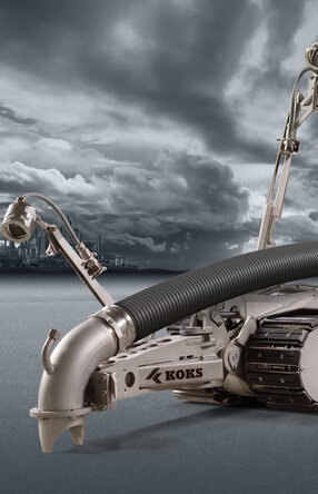 Roboter mit Schlauch, der Richtung Boden zeigt, und Kameras, zur Steuerung durch einen Mitarbeiter. Im Hintergrund ist weit in der Ferne Industrie-Landschaft zu sehen, zusammen mit dunkelgrauen Wolken und Boden.