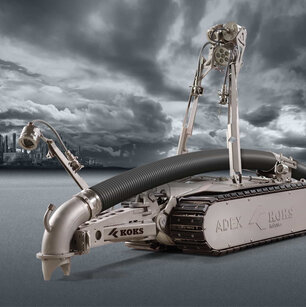 Roboter mit Schlauch, der Richtung Boden zeigt, und Kameras, zur Steuerung durch einen Mitarbeiter. Im Hintergrund ist weit in der Ferne Industrie-Landschaft zu sehen, zusammen mit dunkelgrauen Wolken und Boden.