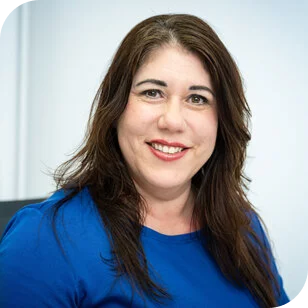 Man sieht eine braunhaarige Frau. Sie lächelt in die Kamera und trägt einen blauen Pullover. Sie st im Vordergrund des Bildes zu erkennen. Der Hintergrund ist unscharf.