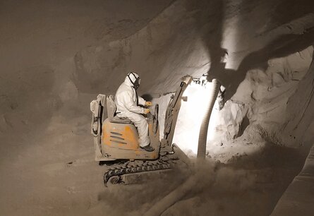 Zu sehen ist ein Arbeiter in Schutzanzug im inneren eines Silos auf einem Bagger ähnliche Fahrzeug an dem ein Schlauch befestigt ist, womit ein Pulver aufgesaugt wird.