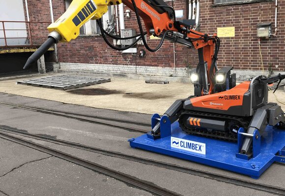 Roboter, orangefarben, mit ausgestrecktem Greifarm, Stift ähnlichem Aufsatz am Ende des Greifarms und kleinen Scheinwerfern, auf einer königsblauen Platte, die über ein Schienensystem auf einem Betriebshof fährt. Im Hintergrund ist eine Backstein-Fassade zu sehen.