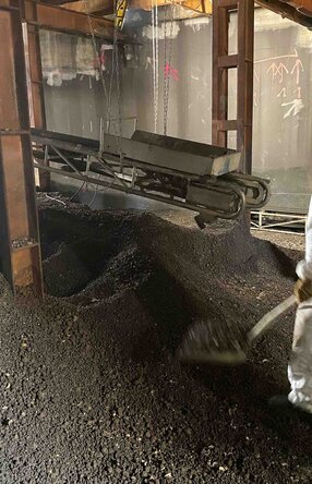 Halle mit einem Haufen an Schutt, darin stehend ein Mitarbeiter in Schutzanzug und Maske mit einer Schaufel, der Schutt auf einen Kasten auf einem Förderband auflädt. Das Förderband hängt zwischen Metallpfeilern und läuft nach hinten aus dem Bild raus.