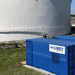 Aufnahme des Inneren eines Tanks, aufgenommen im Tank selbst. Der Boden ist Beige oder Cremefarben, es ragen viele Rohre, oben abgeklebt mit einer schwarzen Folie, aus dem Boden. Im Hintergrund ist ein Verteiler mit weiteren Rohren zu sehen, diese führen aus teilweise aus dem Bild heraus oder verbinden sich mit Ventilen aus dem Boden, die Wand des Tanks ist mit Rost versehen, darüber ist ein Stück freier blauer Himmel zu sehen. Links im Bild befindet sich eine Leiter die aus dem Tanks herausführt und rechts läuft ein Arbeiter den Tankrand entlang.