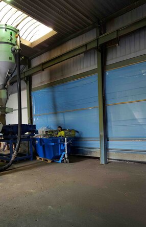 Zu sehen ist eine Halle mit einer relativ großen Siebanlage die von einem Mitarbeiter in neongelber Arbeitskleidung und Schutzhelm bedient wird.