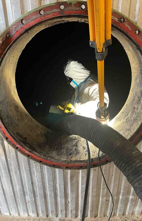 Das Foto zeigt einen von außen Fotografierten Tank mit einer runden Öffnung, im inneren des Tanks steht ein Arbeiter mit Schutzanzug und Maske während er ein Rohr, das von außen kommt, festhält.