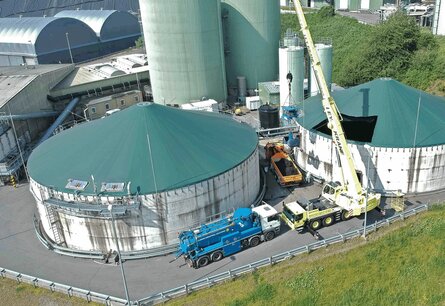 Aufnahme aus der Vogelperspektive, eines grünlichen Fabrikgebäudes, welches aus zwei großen flachen Silos im Vordergrund und zwei schmalen hohen Silos im Hintergrund besteht. Vor und zwischen den Silos stehen unterschiedliche LKWs, einer mit Tank, einer mit offener beladener Ladefläche und einer mit einem Kran der gerade einen Maschine in einen der Tanks hebt. Im Hintergrund sind weitere Fabriken sowieso Wiesen und Wälder zu sehen.