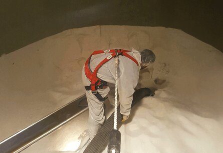 Ein Mitarbeiter in weißem Ganzkörperanzug, Haarnetz und einem roten Geschirr, das an dem ein Seil befestigt ist, steht mittig in einem Tank auf weißem Pulver. Der Mitarbeiter hält mit beiden Händen ein Rohr womit es das Pulver vermutlich aufsaugt, links neben ihm ist ein schwarze Brett um aus dem Tank ein- oder auszusteigen.