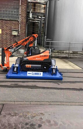 Roboter, orangefarben, mit gesenktem Greifarm stehend auf einer königsblauen Platte die über ein Schienensystem auf einem Betriebshof fährt. Im Hintergrund sind eine Backstein-Fassade und Silos oder Tanks zu sehen.